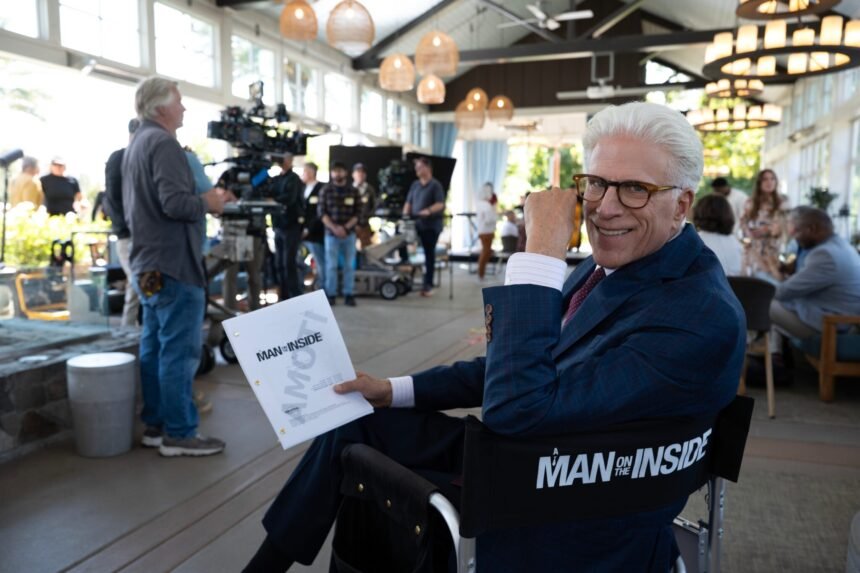 "Man with white hair and glasses sitting in a director’s chair holding a script on a film set, with crew, cameras, and lighting equipment visible in a bright indoor setting."