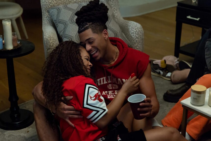"Two people sit on the floor in a cozy living room, laughing and embracing, surrounded by candles and drinks during a casual gathering."
