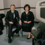 "Two people in formal attire sit side by side in a plain office-like room, holding hands, being filmed by a video camera during an interview or press conference."