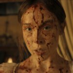 "A young woman with long hair, dressed in light clothing, has her face and upper chest covered in blood, standing indoors with dim warm lighting and blurred curtains and furnishings in the background."