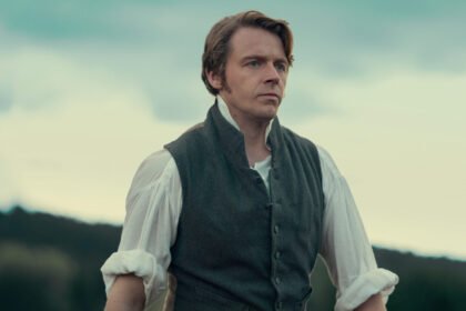 "A man in historical clothing stands outdoors with a serious expression, wearing a vest over a white shirt, with hills and trees in the blurred background under a cloudy sky."