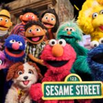 "Group of colorful Sesame Street puppet characters outdoors smiling together, holding a Sesame Street sign, with brownstone buildings and greenery in the background."
