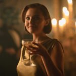 "Woman in a gold dress holding a drink at an elegant, dimly lit party with candlelight in the background, creating a warm, sophisticated atmosphere."